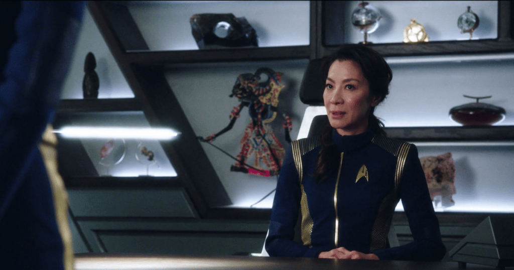 Captain Georgiou sitting at her ready room desk; the shelves behind her hold decorative objects including a Malaysian wayang kulit shadow puppet.