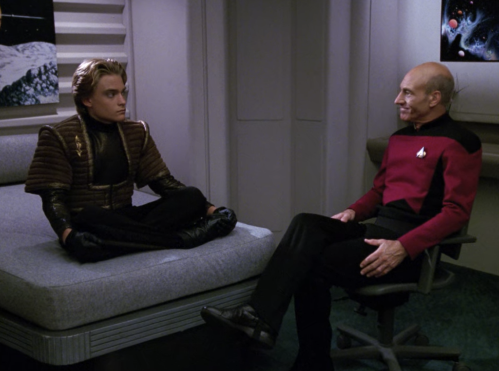 Jono sitting cross-legged on a bed, talking to Picard