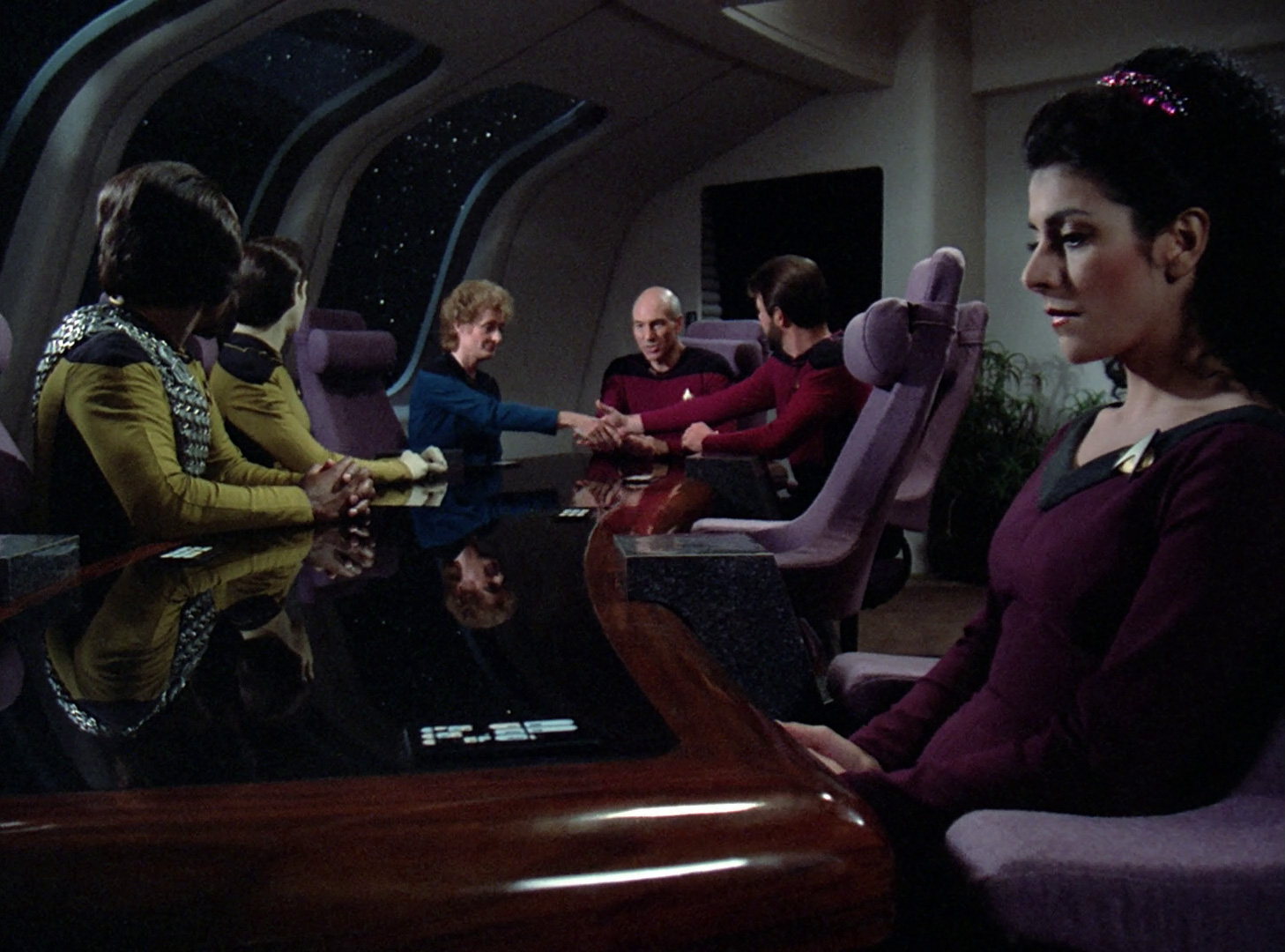 Troi sits by herself at the end of the table while the others discuss her pregnancy